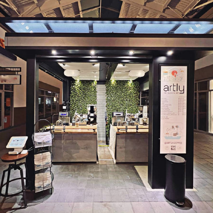 A freestanding Artly location has come to life. Greenery lines the back wall. The black paneling, wood counters, and bright lighting make it an inviting place for people to get coffee.