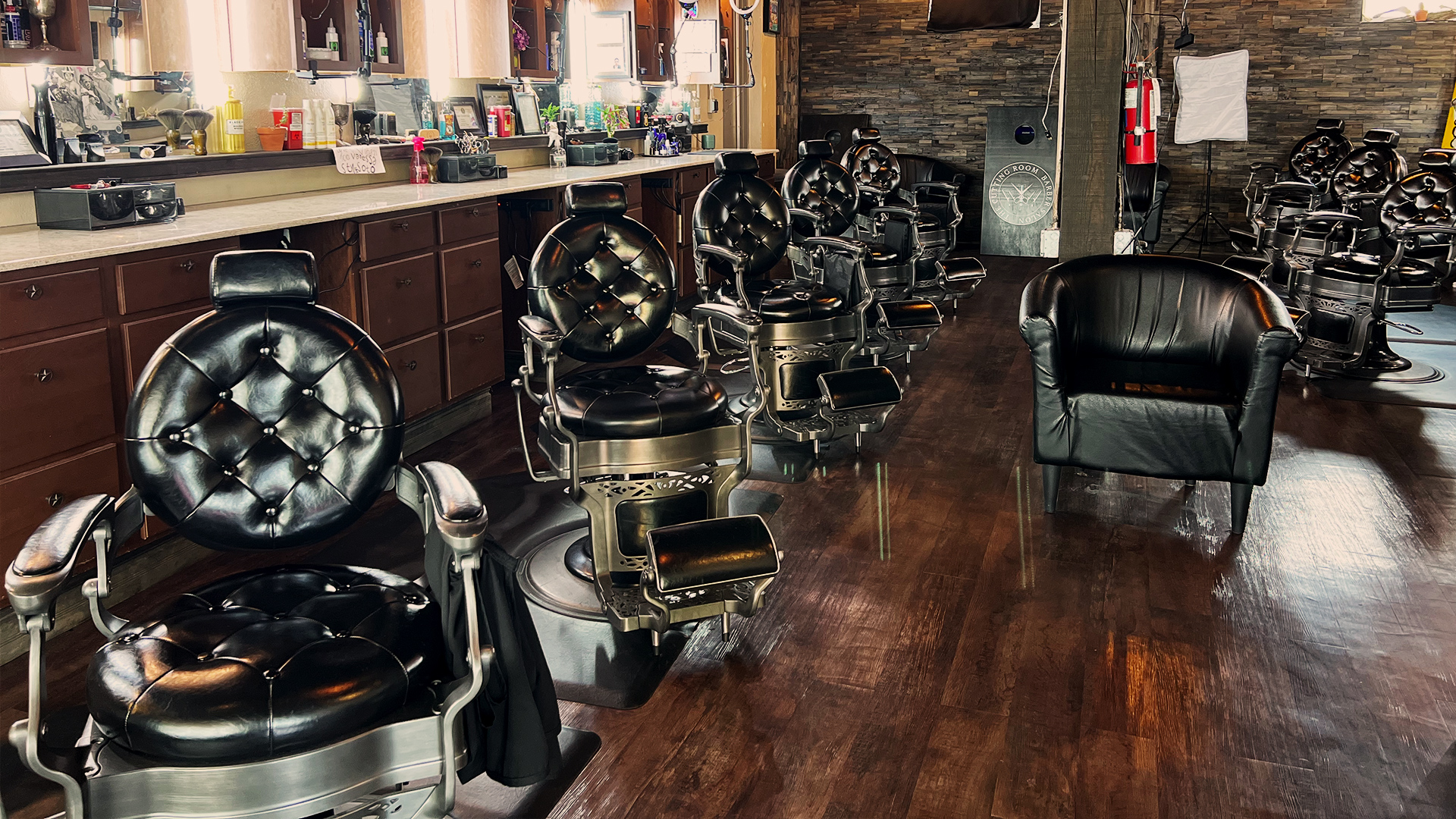 Black leather barber chairs are in two rows with another black leather lounge chair in the middle. Dark hardwood floor and dark cabinets serve a stark contrast to the white counters and lights around the mirrors.