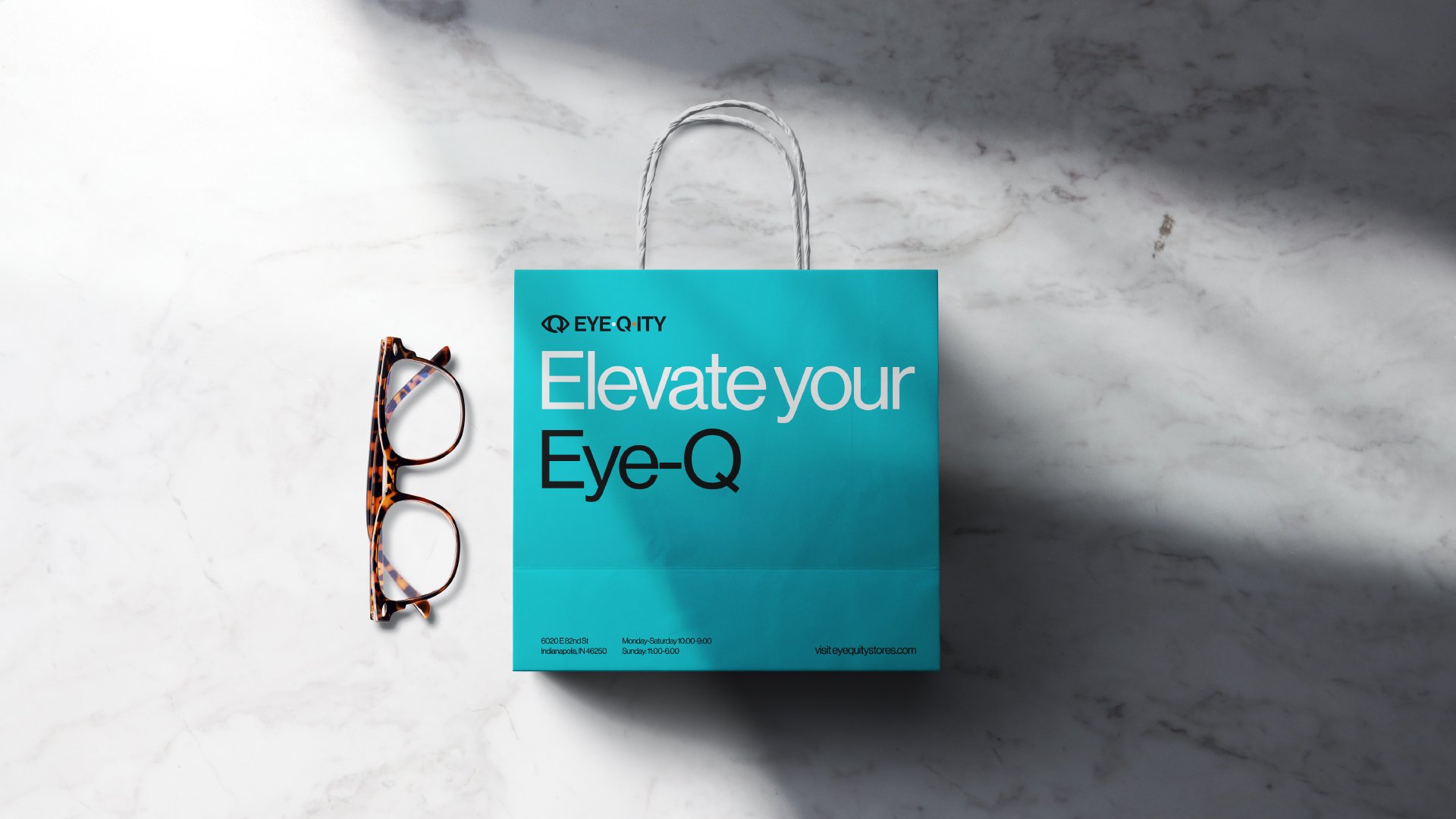 Glasses sit next to an EYEQ.ITY bag. It says, “Elevate your Eye-Q” with the logo above it and the contact information and address below it. Both the bag and the glasses are on top of a marble surface.