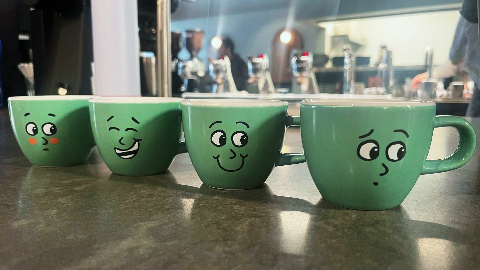 Four teal mugs featuring different Collin expressions sit side by side. The coffee shop is in the background.