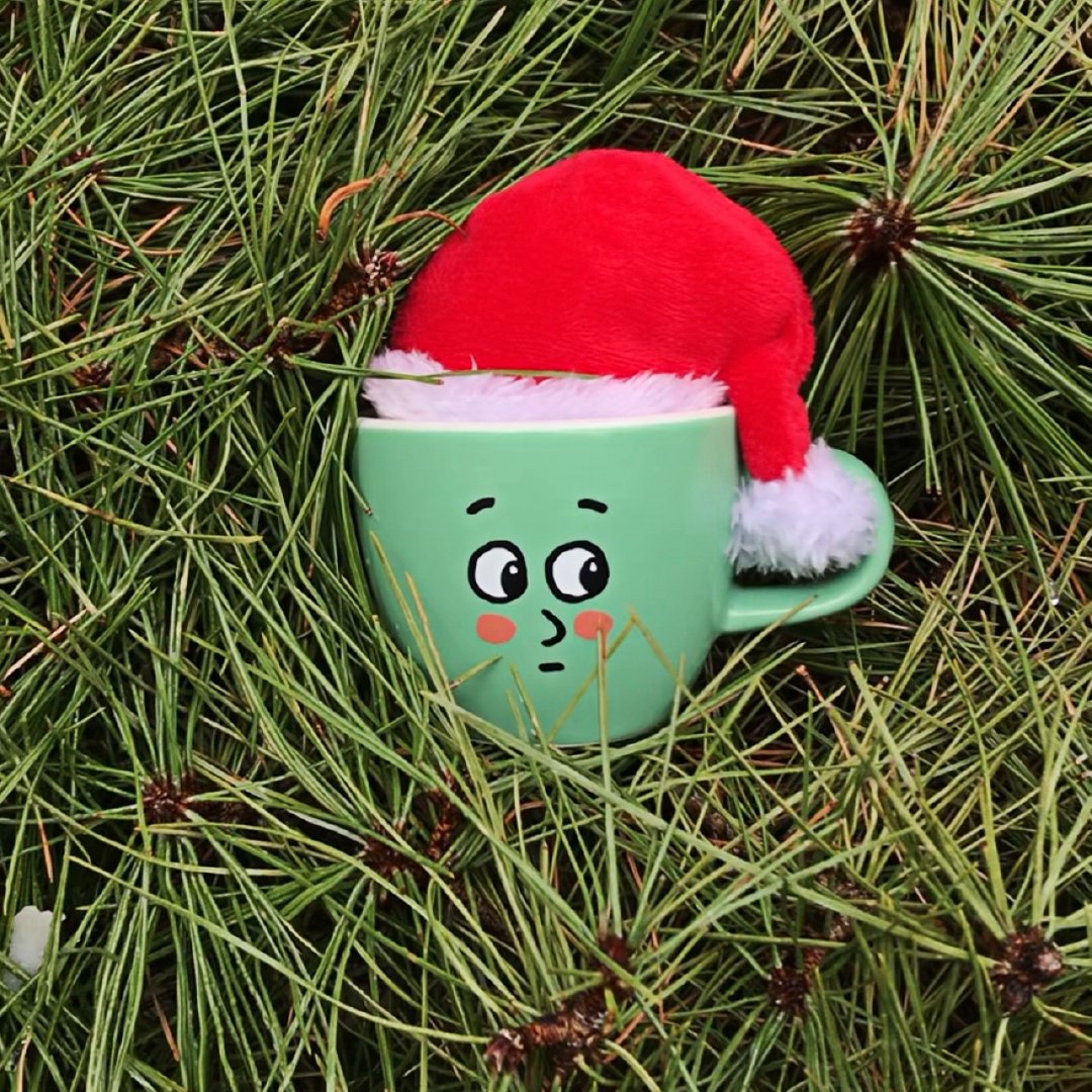 A teal coffee mug with the face of “embarrassed Collin” fits snug among branches and pine needles while wearing a Santa hat.