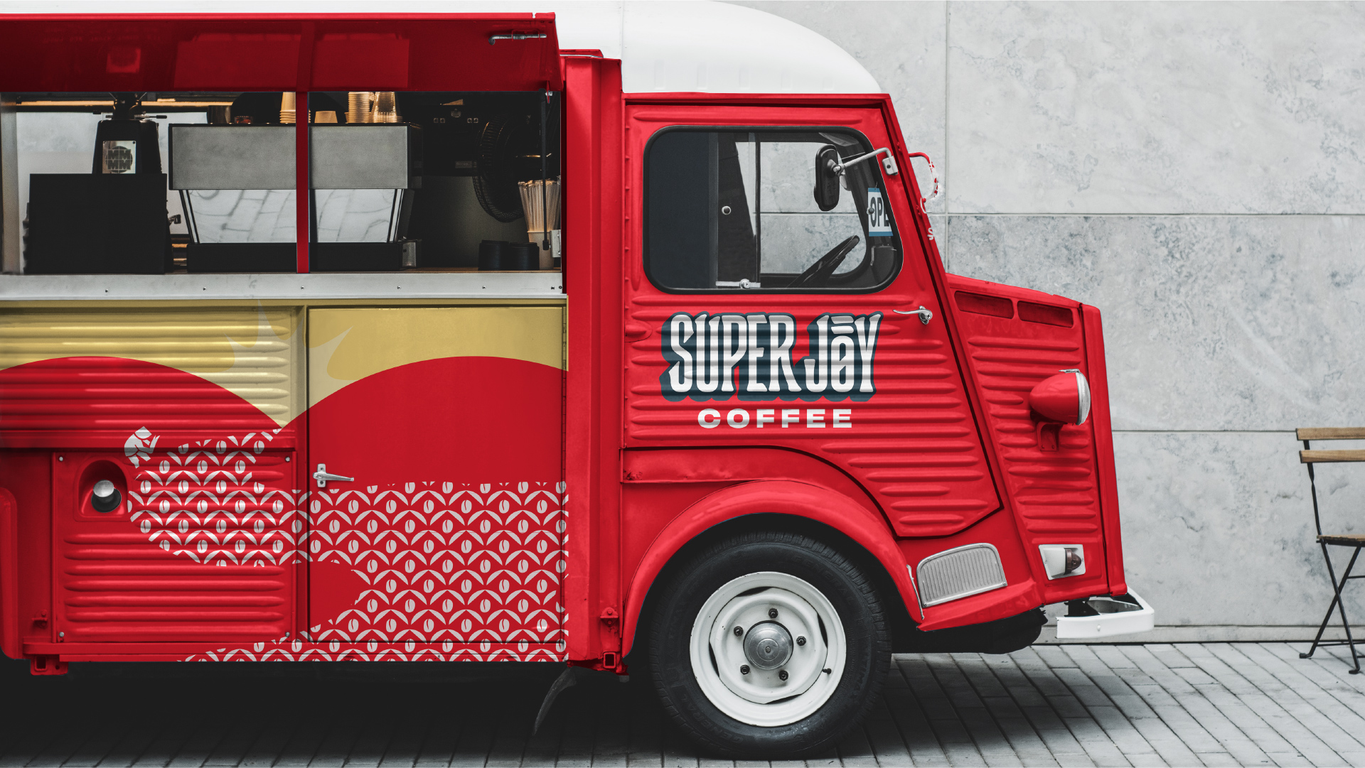 A Super Joy mobile cafe is a striking red truck with the logo on the passenger door. The side panel shows rolling hills with a yellow rising sun. There are illustrated beans among the hills.