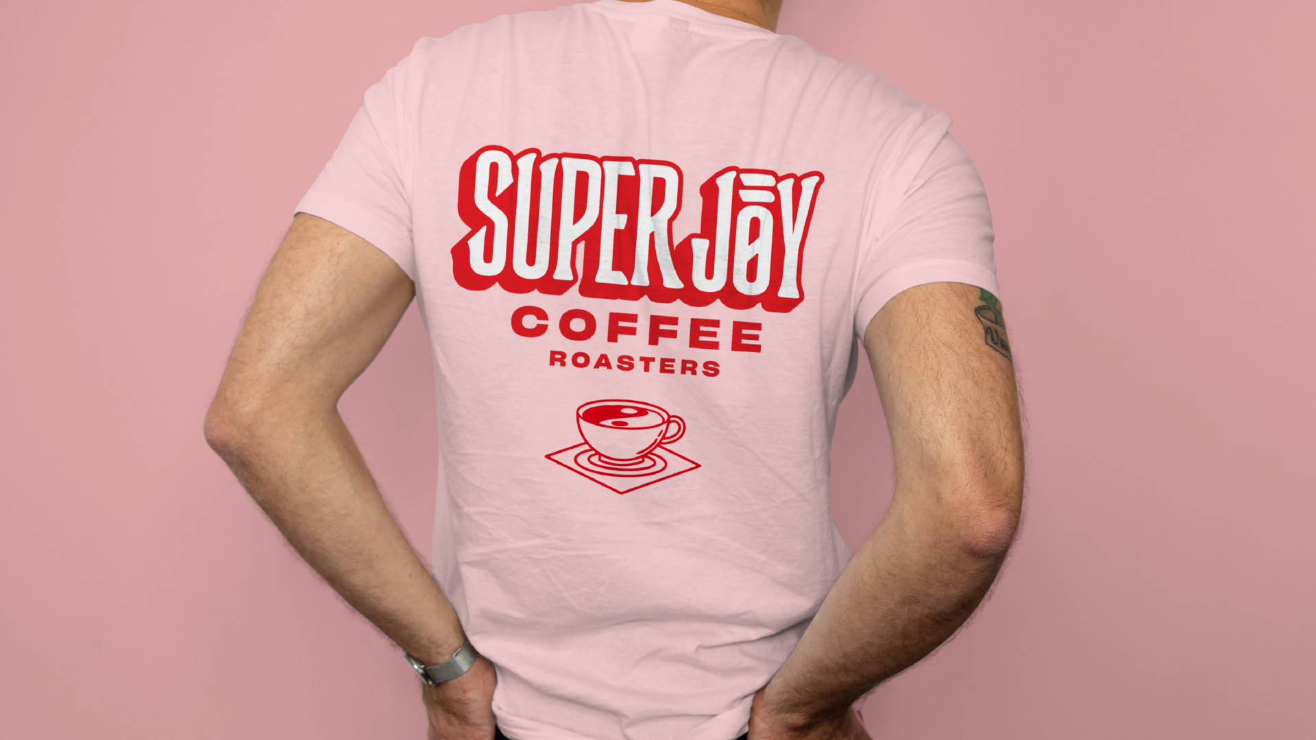 A person wears a short-sleeve pink Super Joy shirt. The back has the Super Joy logo with a coffee cup below it. In the coffee cup is the yin and yang symbol.