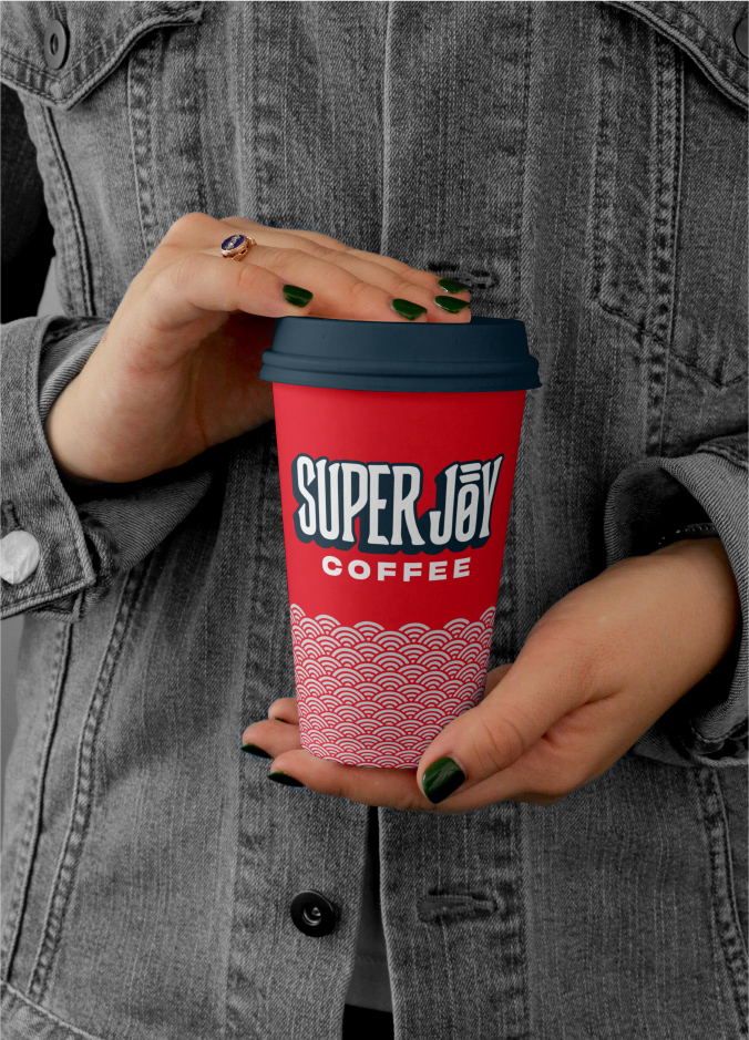 Someone with a gray jean jacket and painted nails holds a Super Joy to-go cup. It’s red with the Super Joy Coffee logo and a white circular design on the bottom half of the cup.